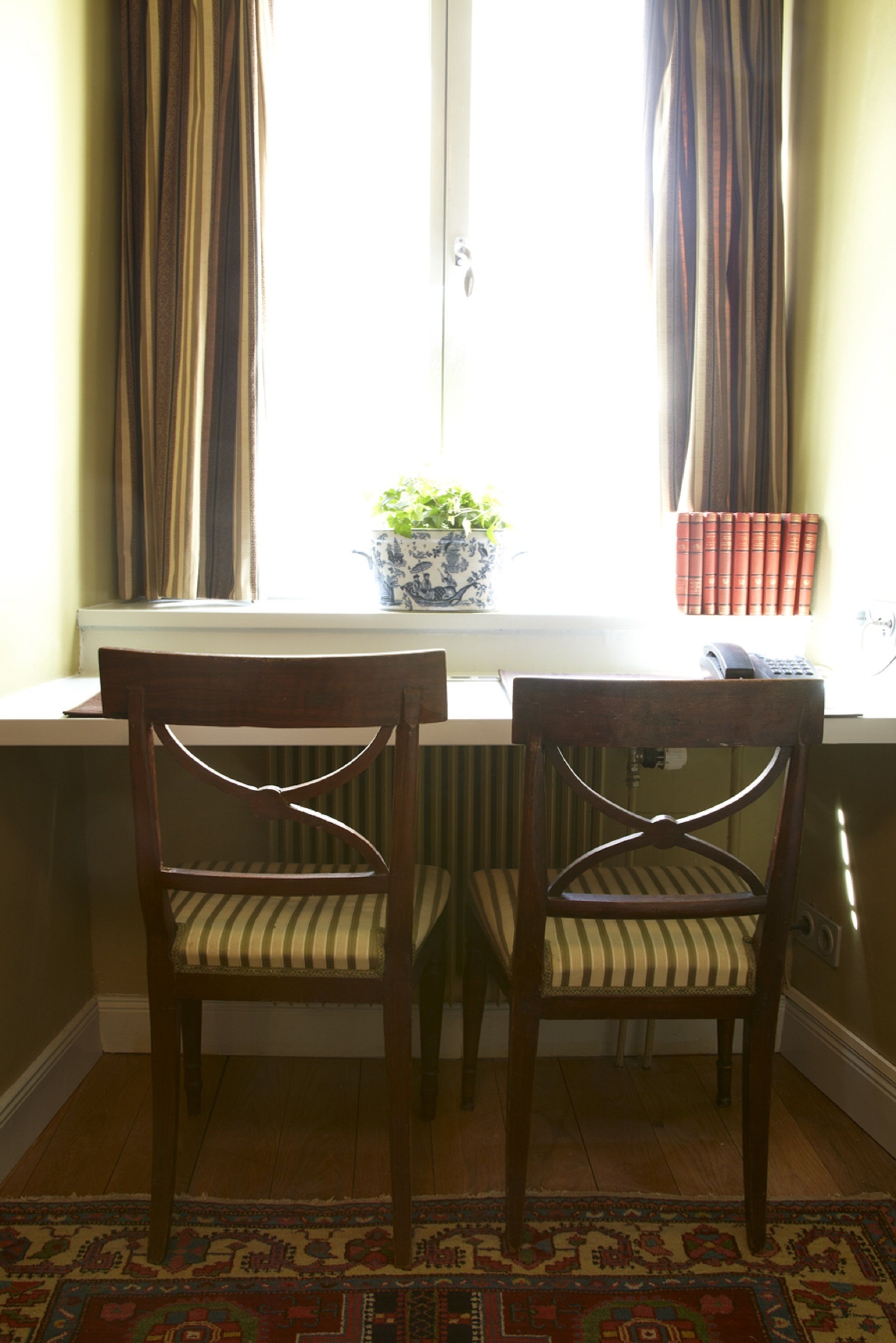 Two wooden chairs with striped cushions at a window sill, a potted plant, and a row of books on the windowsill.