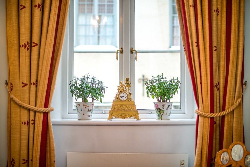 Une fenêtre avec des rideaux, deux plantes en pot et une horloge décorative posée sur un radiateur.
