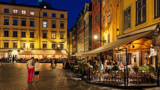 A cobblestone square with outdoor seating at a café, surrounded by historic buildings with multiple stories and a church spire in the background.