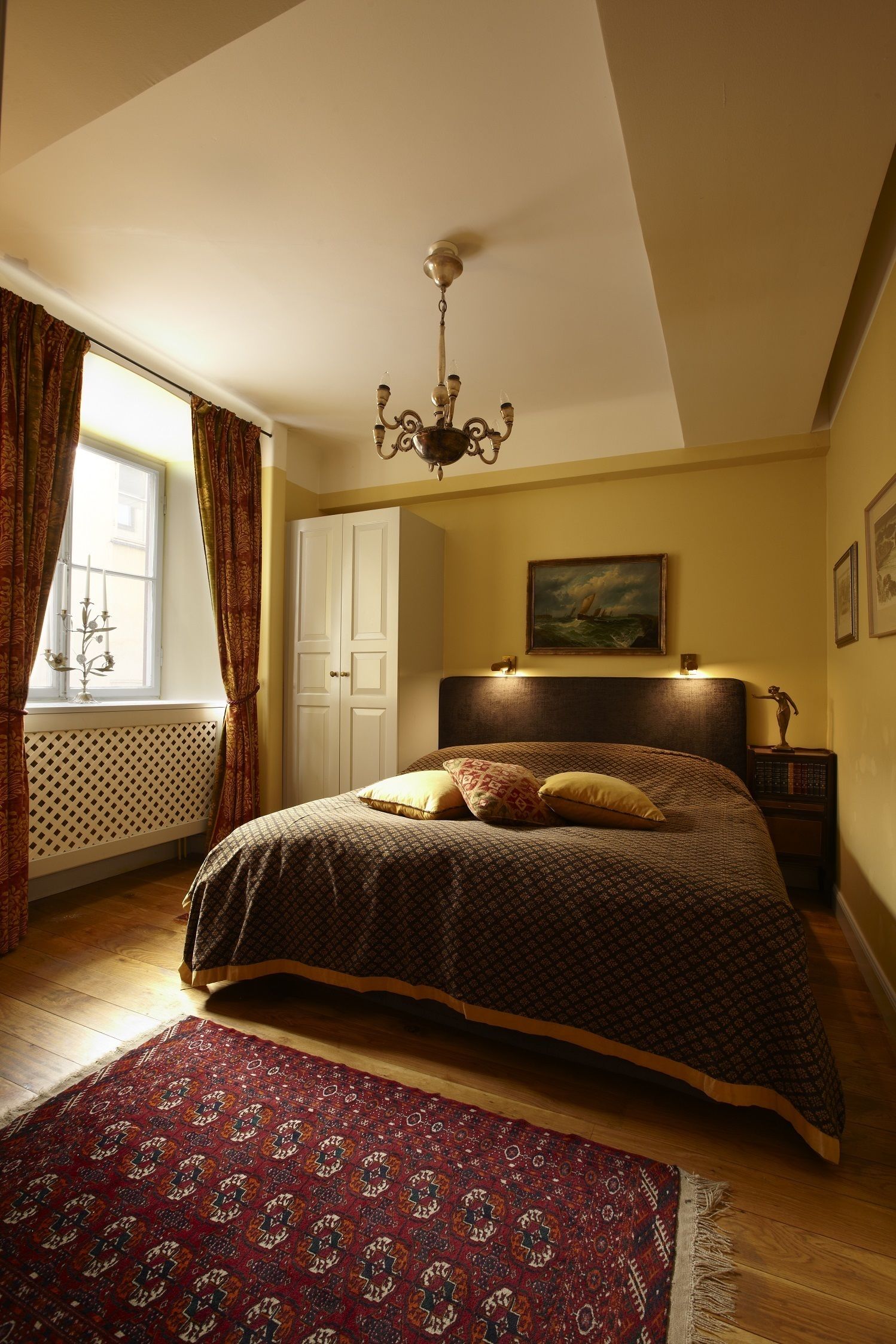 Ein Schlafzimmer mit einem Bett, einer Kandelaber, einem Gemälde, einem Kleiderschrank und einem gemusterten Teppich.