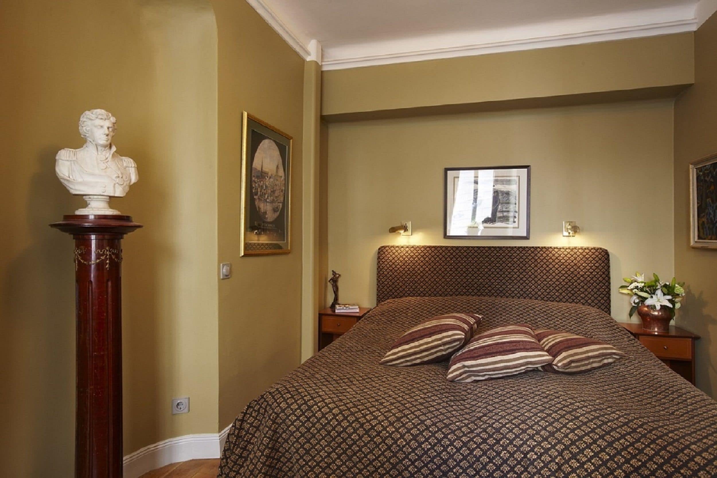 A bedroom with a patterned bedspread, a bust sculpture on a pedestal, and framed pictures on the wall.