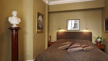 Een slaapkamer met een bed met een patroon, een borstbeeld op een voetstuk, en ingelijste foto's aan de muur.