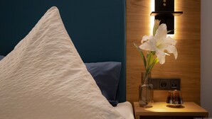 A neatly made bed with a white pillow and a blue headboard. A bedside table with a vase of white lilies and a small lamp is visible. - Landhotel Zur Guten Einkehr (Doberschau-Gaussig)