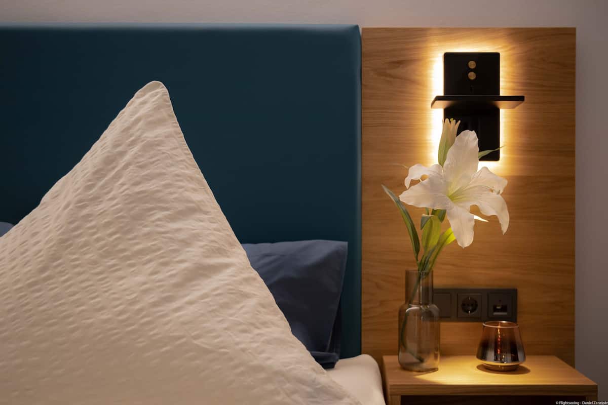 Un lit bien fait, avec un oreiller blanc et une tête de lit bleue. On aperçoit une table de chevet sur laquelle se trouve un vase rempli de lys blancs et une petite lampe.