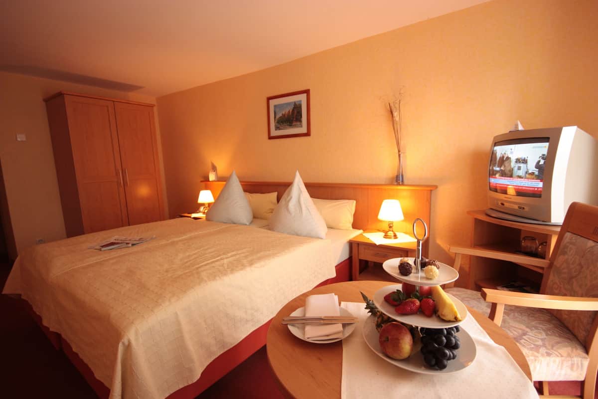 Une chambre d’hôtel avec un grand lit, des tables de chevet avec des lampes, une télévision et un plateau de petit-déjeuner avec des fruits et des viennoiseries.