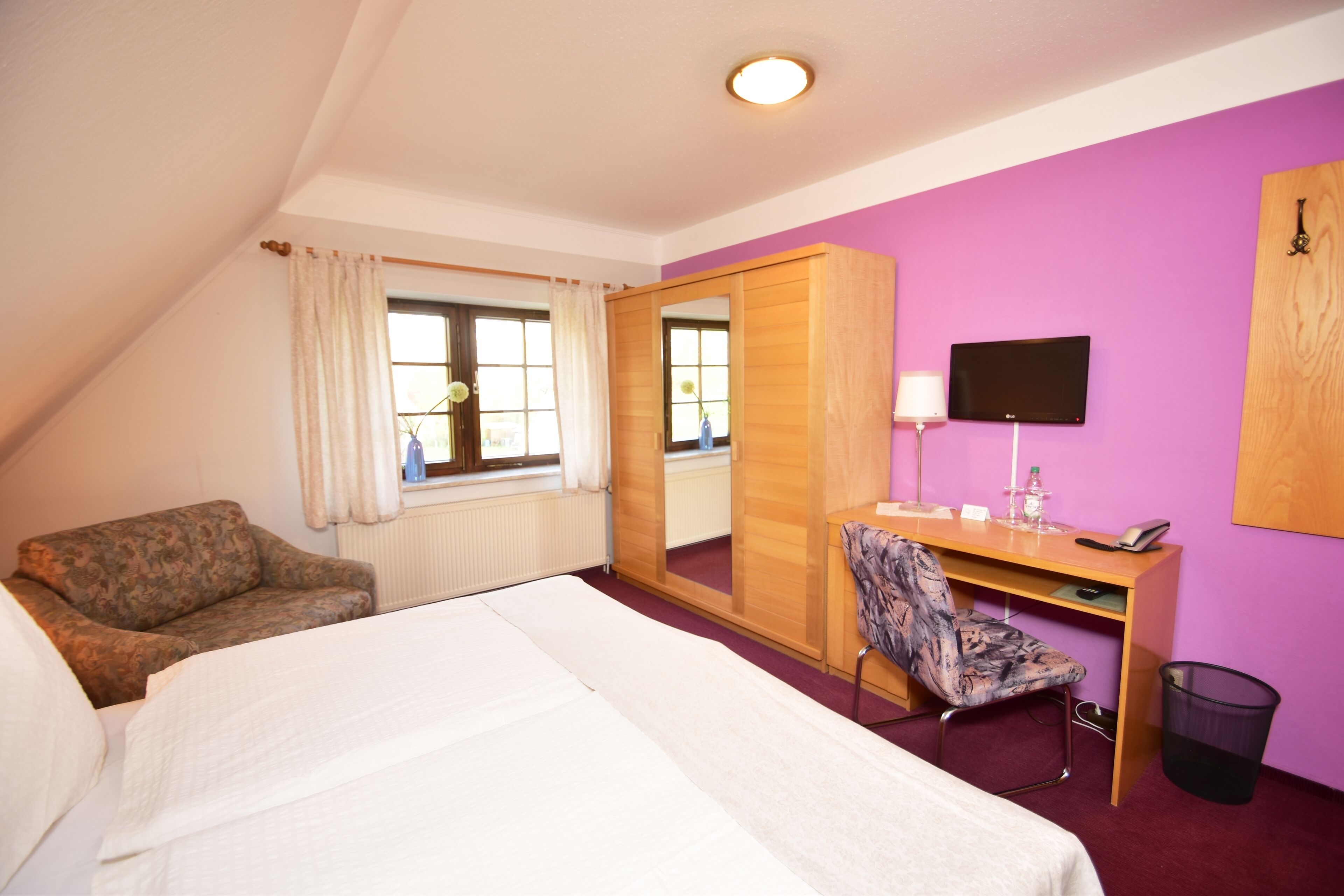 A hotel room with a bed, a desk with a chair, a TV, a mirror, and a window with curtains.