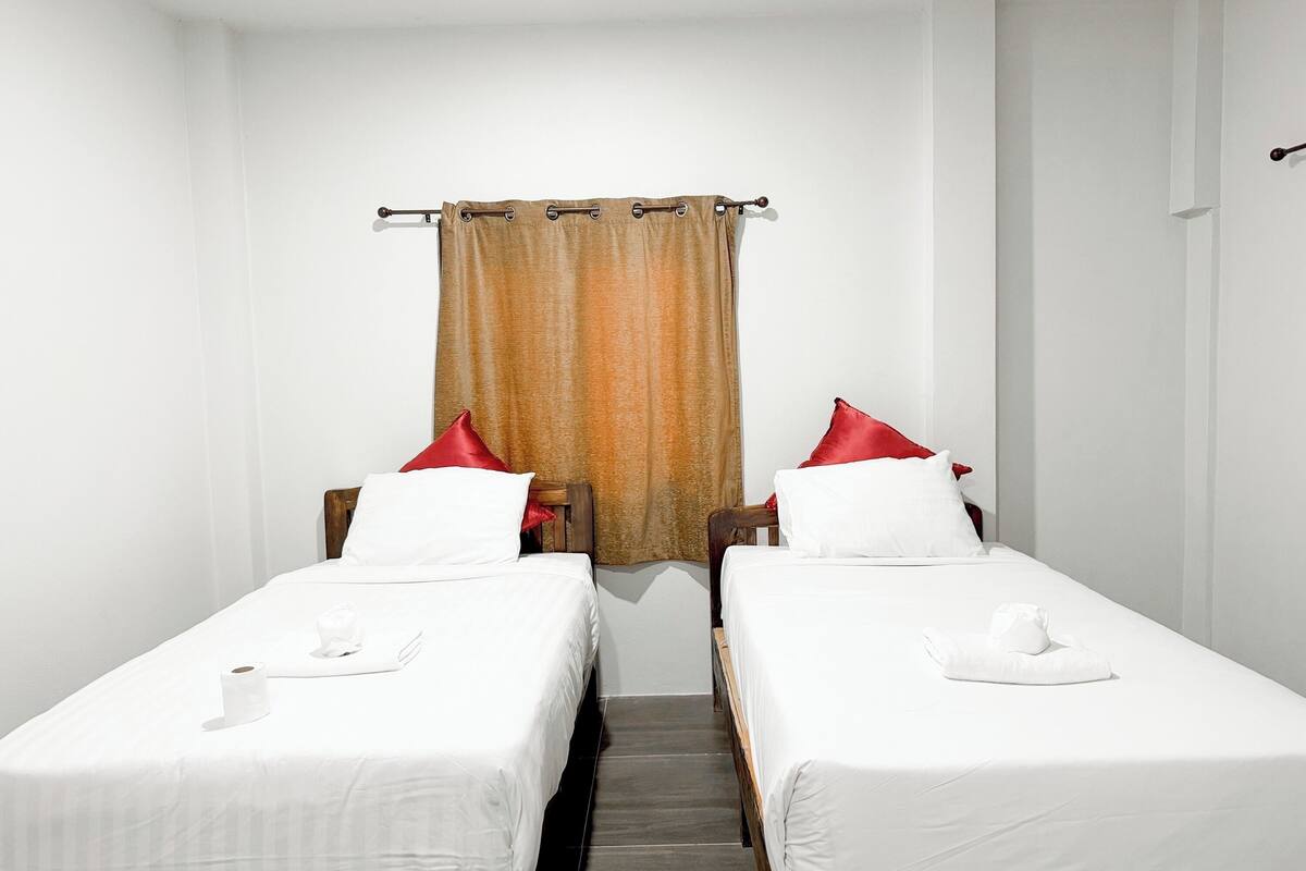 A room with two single beds, each with white bedding and red pillows. There is a curtain with a brownish pattern, and the walls are white.