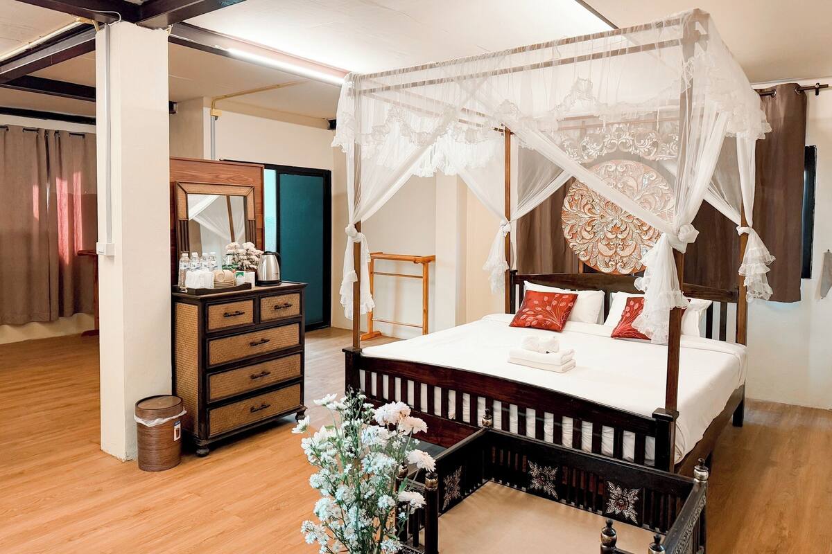 A four-poster canopy bed with a wooden frame and white bedding, a wooden dresser with drawers, a mirror, and a small table with toiletries.