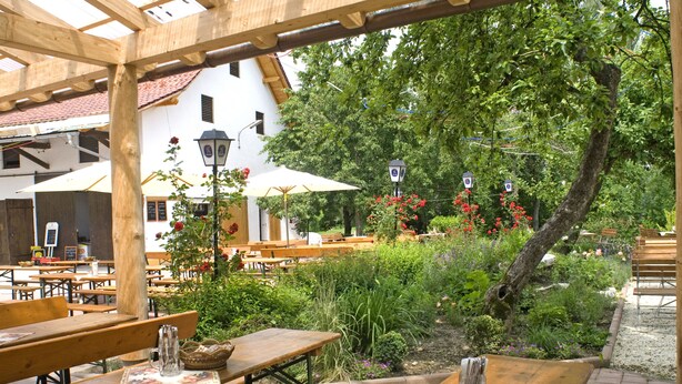 An outdoor seating area with wooden tables and benches, surrounded by lush greenery and flowers. - Hotel zum Fischerwirt (Ried)