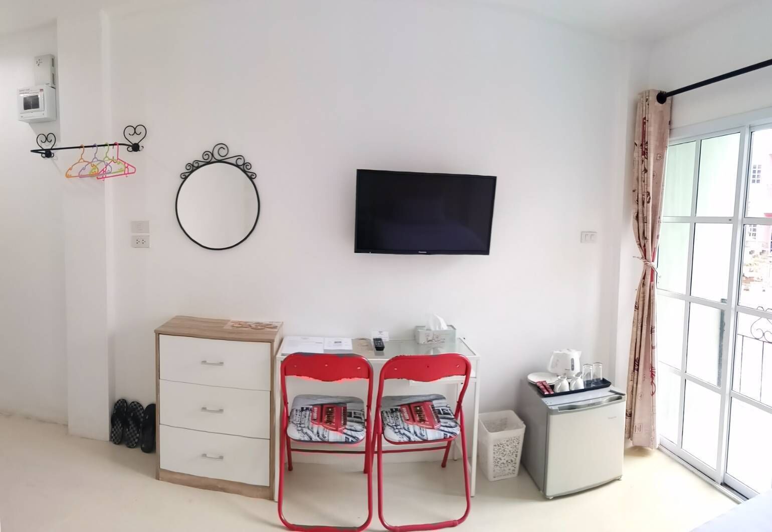 A small, bright room with a desk, two red chairs, a television, and a window with curtains.