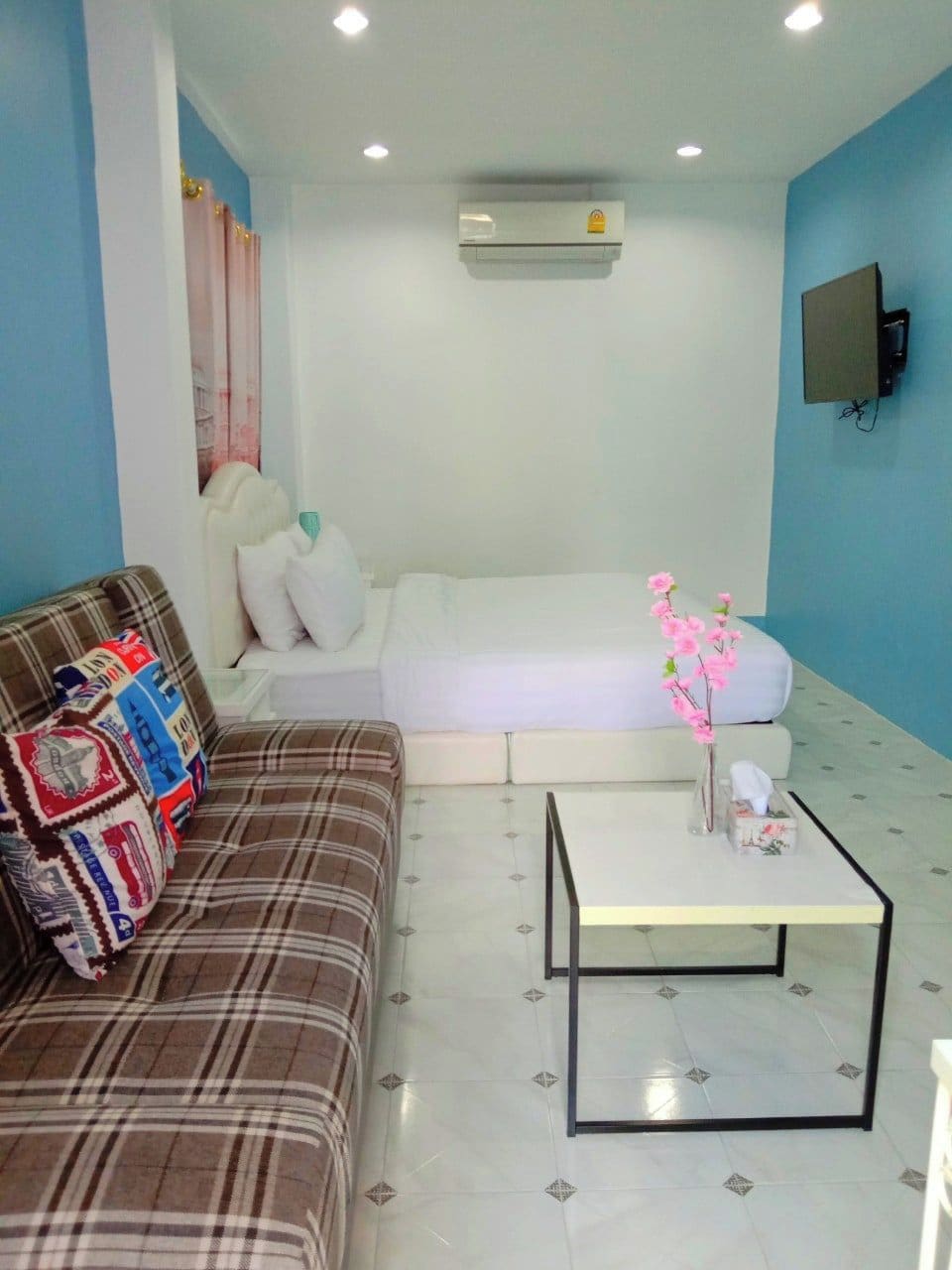 A room with a brown plaid sofa, a white sofa, a small white table, and an air conditioning unit on the wall.