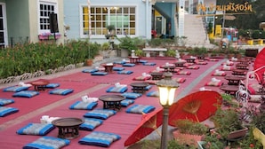 An outdoor seating area with tables and blue cushions, surrounded by potted plants and a red umbrella.