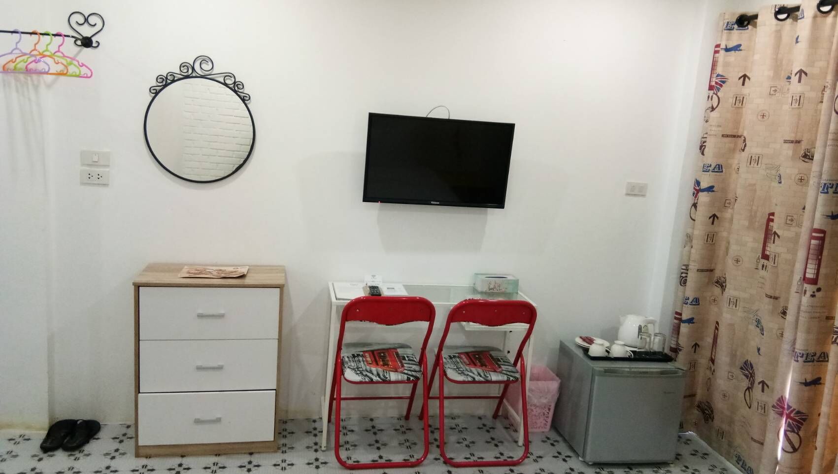 Une petite pièce comprenant un bureau, deux chaises rouges, un téléviseur, un miroir et un rideau orné d’un motif nautique.
