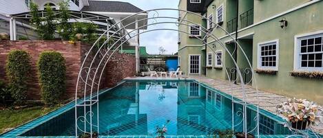 A swimming pool with a white metal arch, surrounded by a white fence, and adjacent to a building with a green facade and multiple windows.