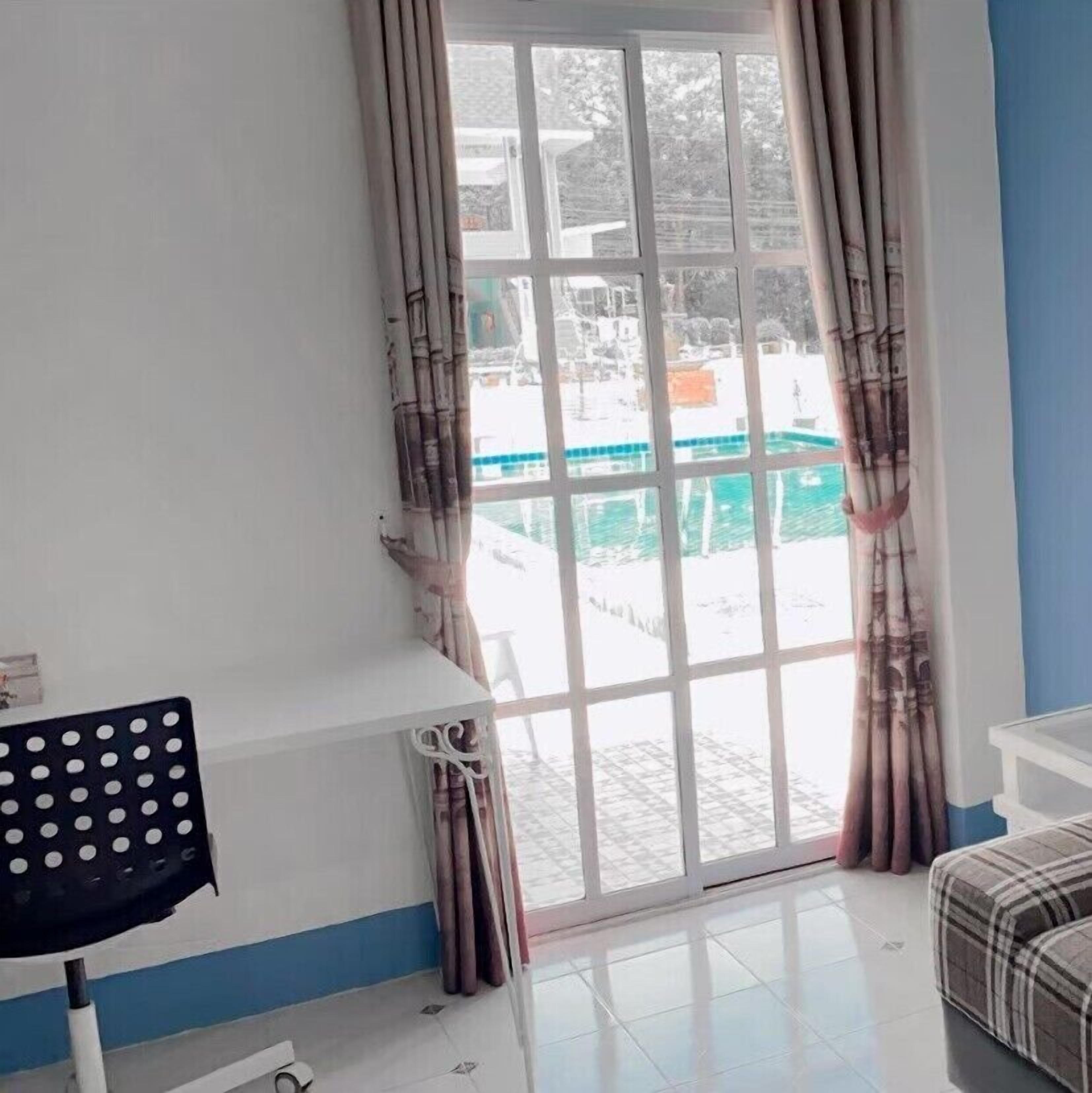 A room with a white desk, a chair, a sofa, and a sliding glass door leading to an outdoor area with a pool.