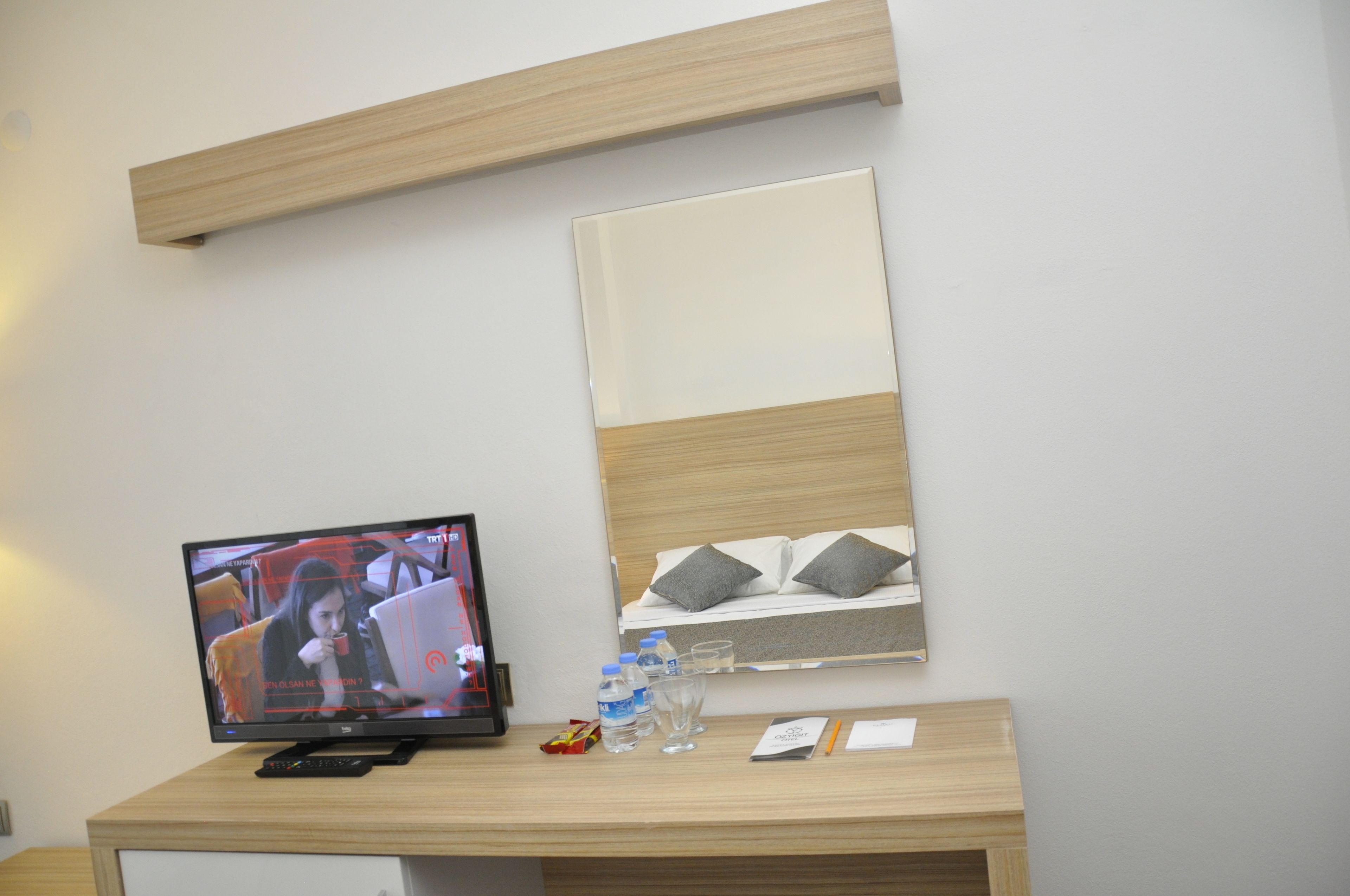 Quarto de hotel com escrivaninha de madeira, televisão de tela plana, cama com almofadas e mesa de cabeceira com garrafa de água, copos e controle remoto.