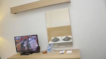 Quarto de hotel com escrivaninha de madeira, televisão de tela plana, cama com almofadas e mesa de cabeceira com garrafa de água, copos e controle remoto.