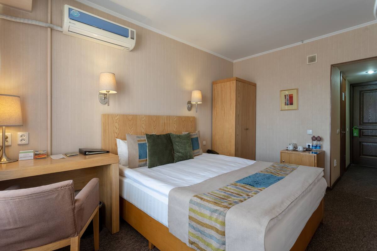 Habitación de hotel con cama, escritorio, silla, armario, mesita de noche, lámpara y unidad de aire acondicionado.