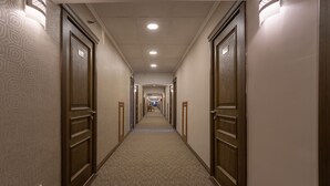 Un lungo e stretto corridoio d'hotel con porte in legno su entrambi i lati, pavimento rivestito di moquette e luci a incasso nel soffitto.