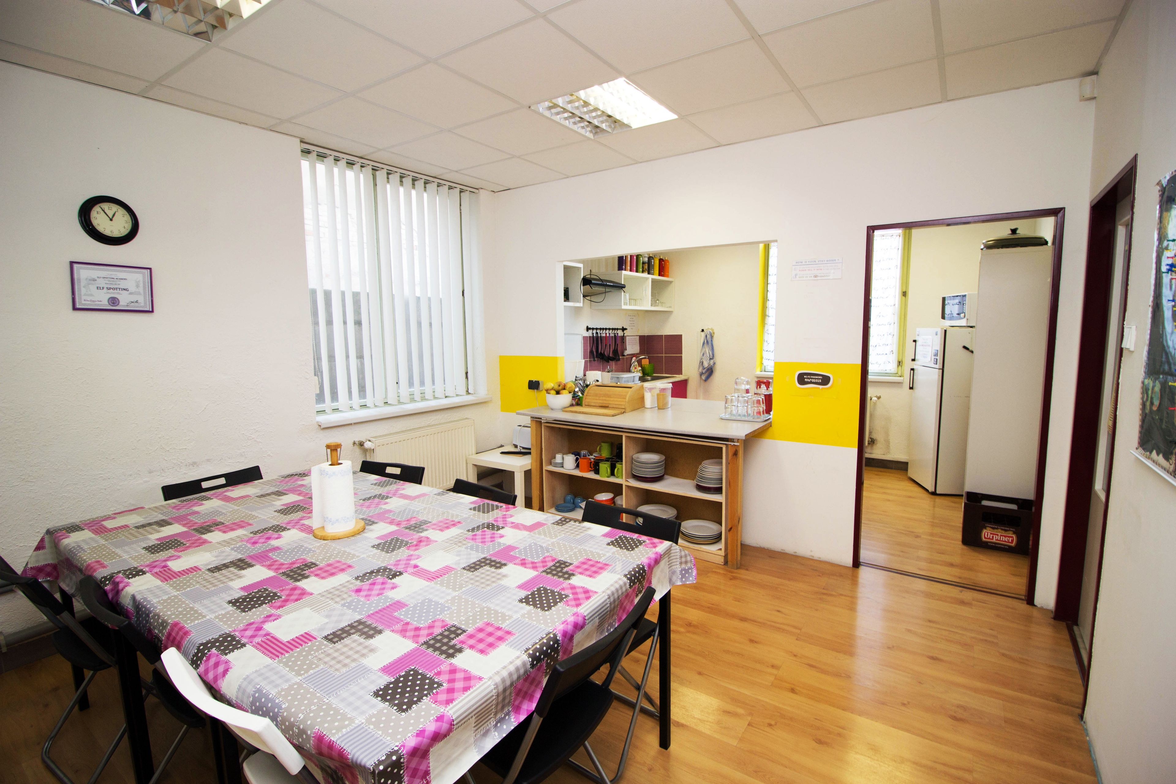 A dining area with a table covered in a quilt, chairs, a clock on the wall, and a kitchen area in the background.