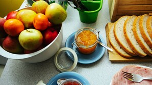 Una encimera de cocina con un bol de manzanas y naranjas, un tarro de mermelada, rebanadas de pan y un plato de jamón.