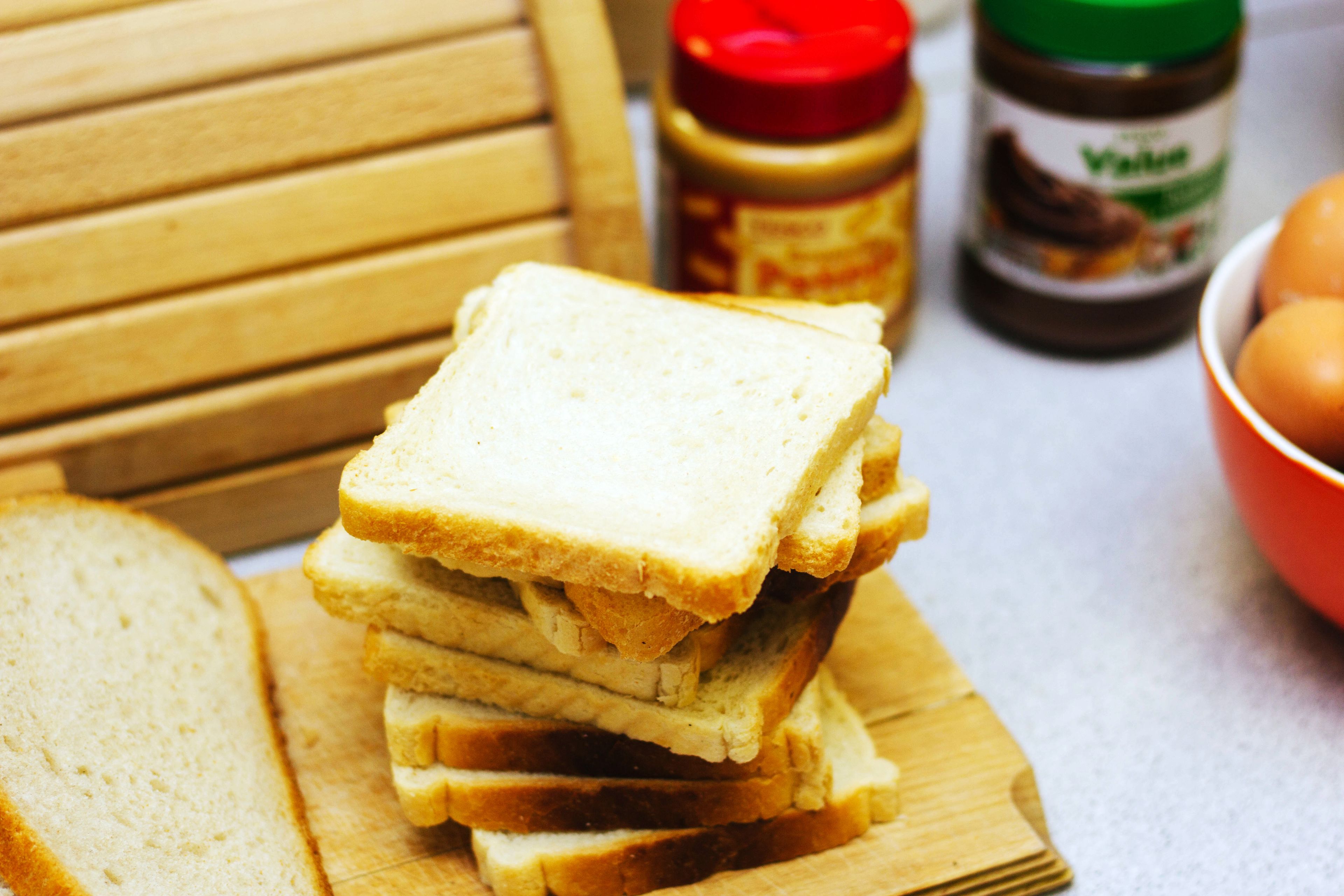 A stack of white bread slices on a wooden cutting board, with a jar of peanut butter and a bowl of eggs in the background.