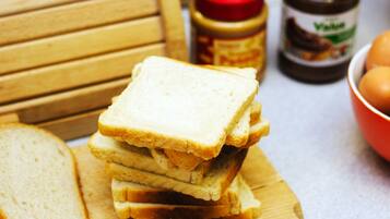A stack of white bread slices on a wooden cutting board, with a jar of peanut butter and a bowl of eggs in the background.