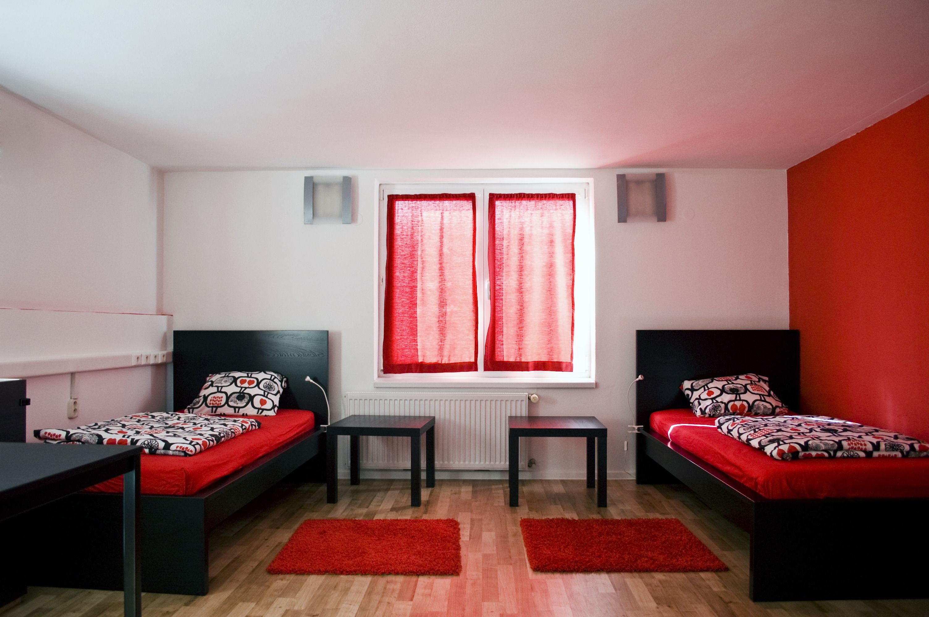 A room with two single beds, a small table, two chairs, a radiator, and a window with red curtains.