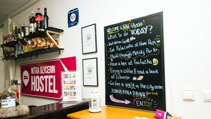 Interior de un hostal con una pizarra que anuncia actividades, un cartel de Nitra Glycerin Hostel y estantes con botellas y aperitivos.