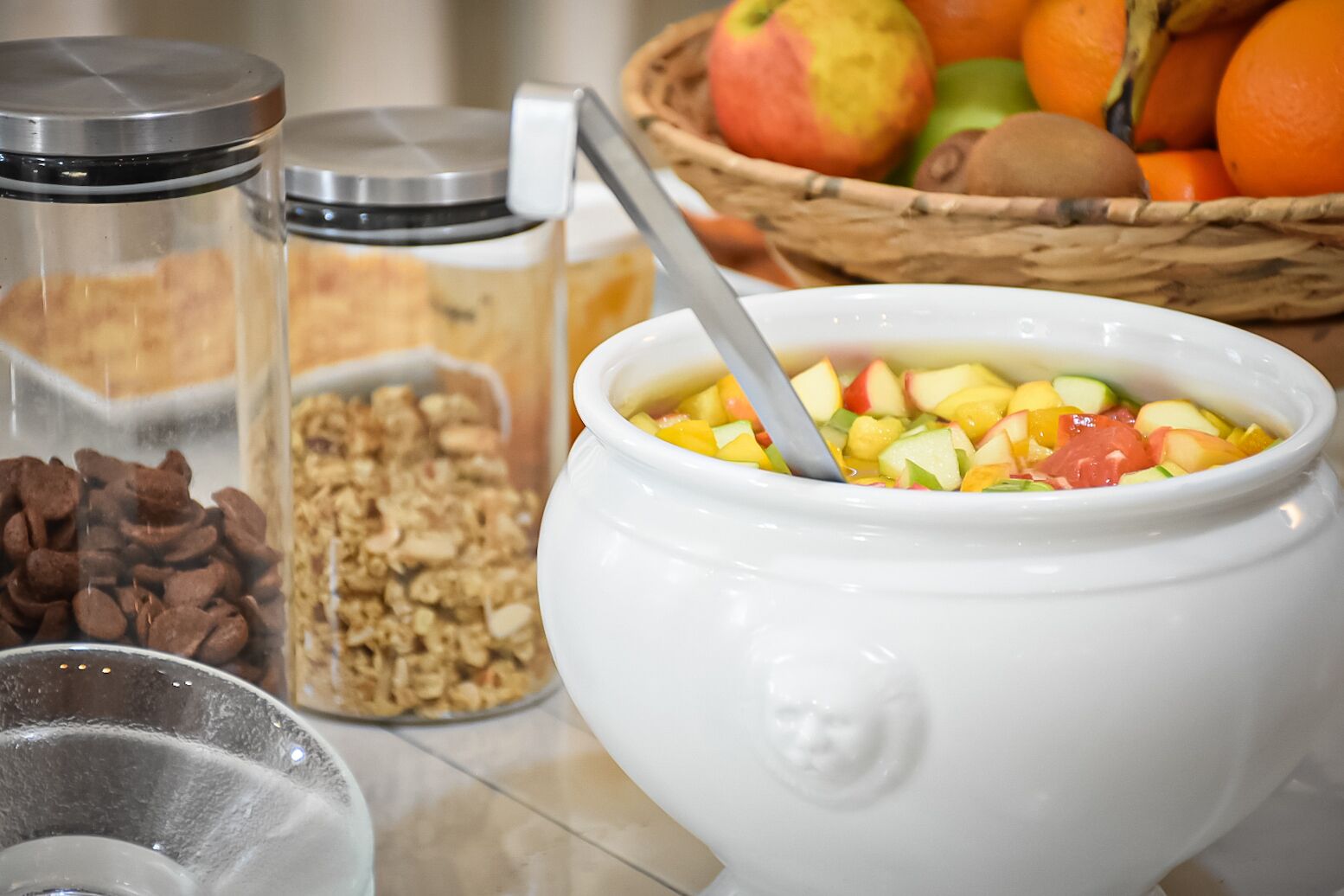 Un tazón de ensalada de frutas con una cuchara, rodeado de recipientes de granola y nueces.