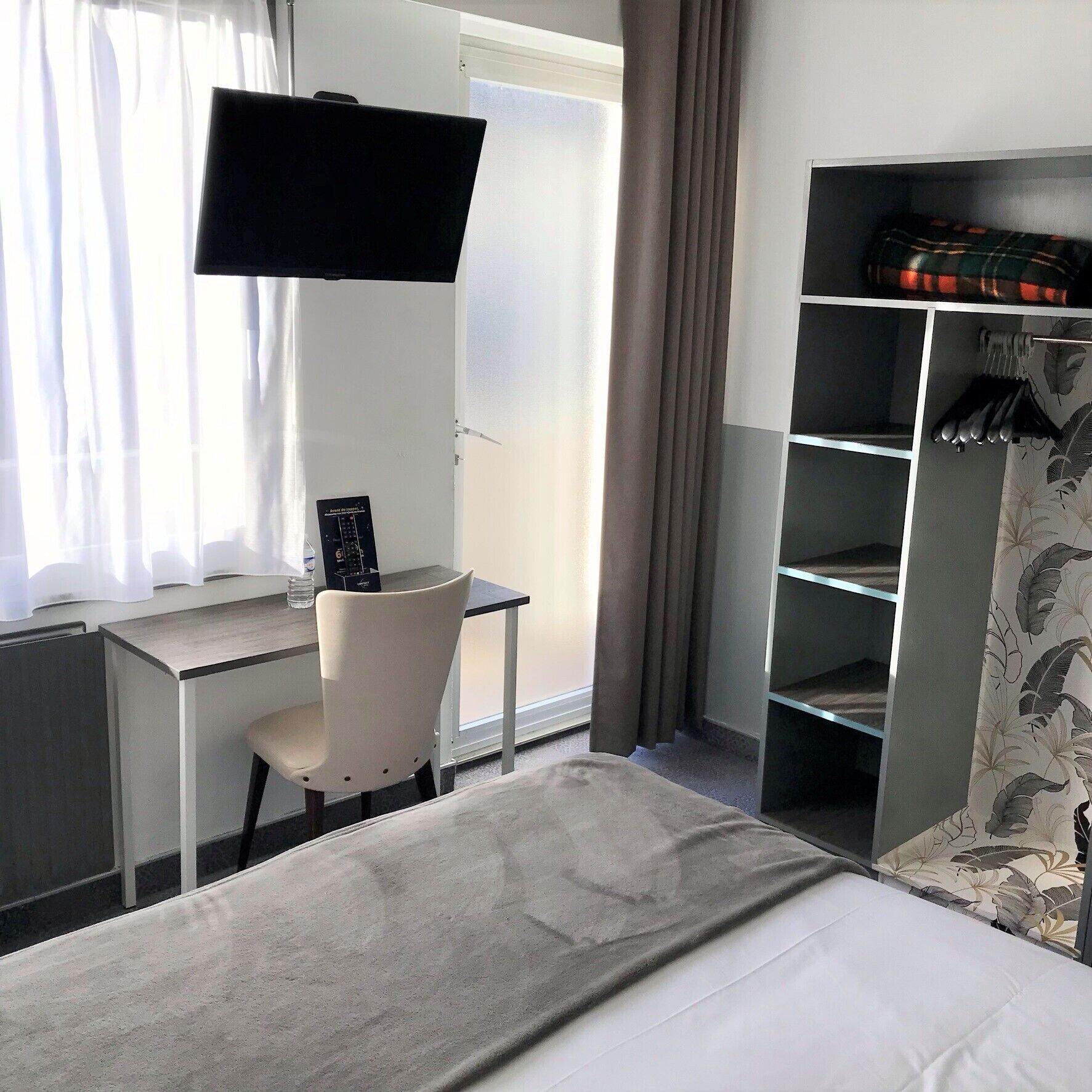 Habitación de hotel con cama, escritorio, silla, televisor montado en la pared y armario con cortina floral.