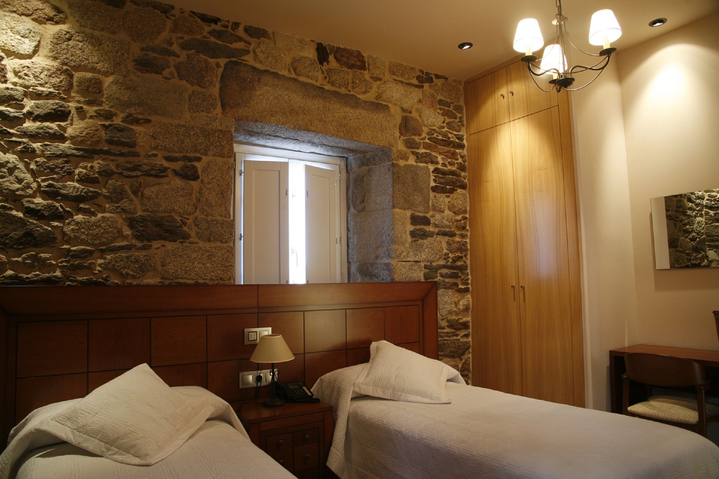 Un dormitorio con dos camas, cabeceros de madera, mesitas de noche con lámparas y una pared de piedra.