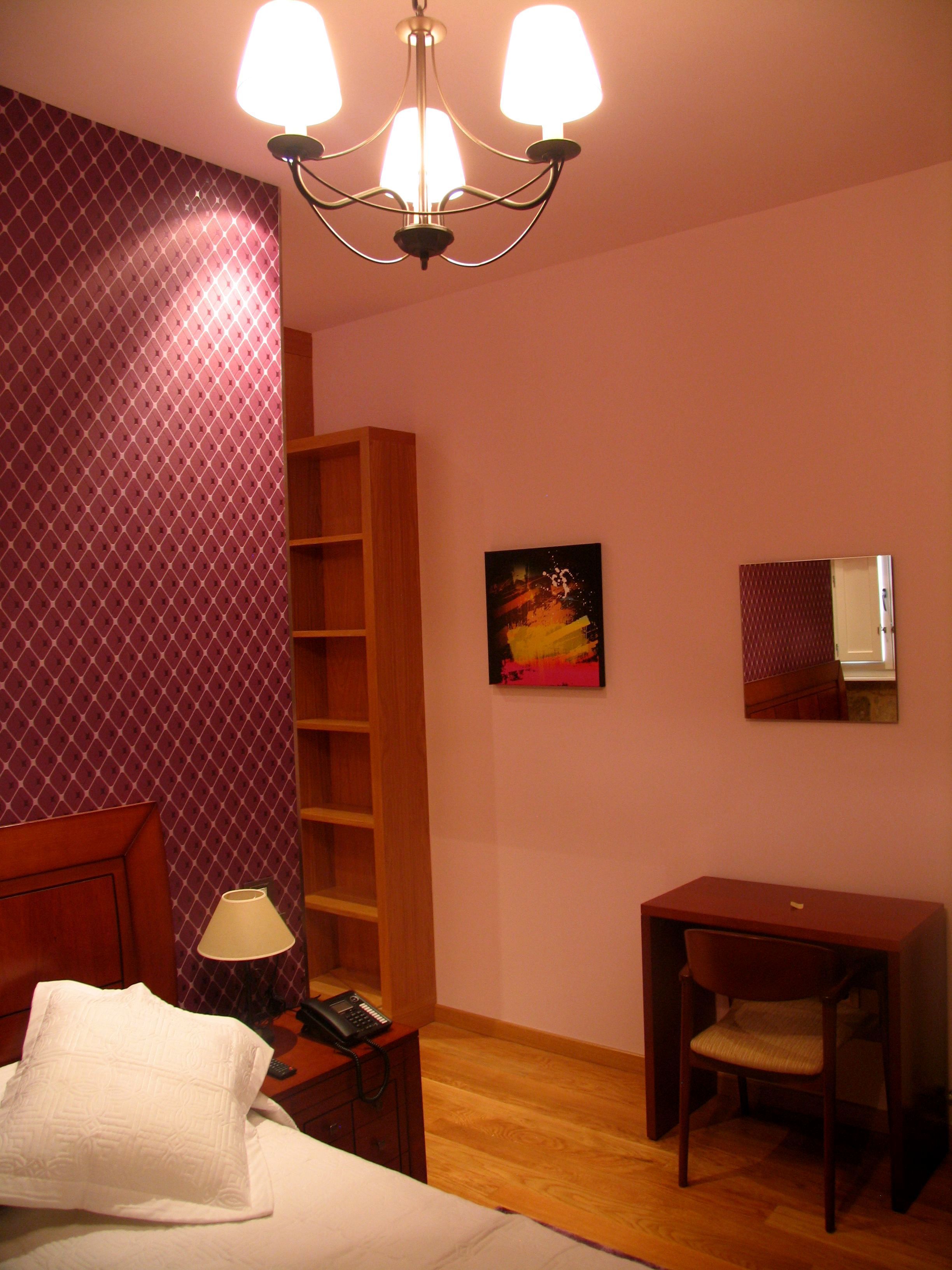 A hotel room with a bed, a desk, a chair, a lamp, a telephone, a bookshelf, a painting, and a mirror.