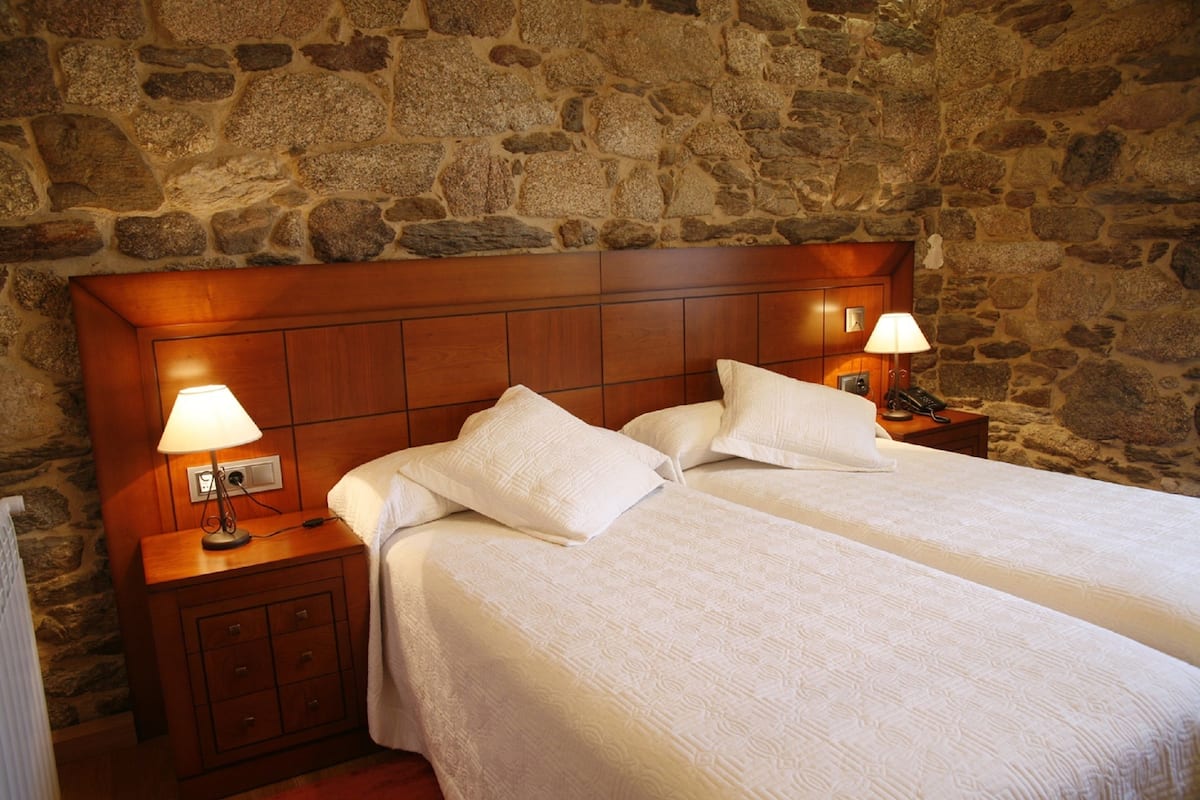 Cama de casal com lençóis brancos num quarto com paredes de pedra e cabeceira de madeira.