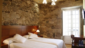 Une chambre avec un mur en pierre, une tête de lit en bois, deux lits avec du linge de lit blanc, une table de chevet avec une lampe et un bureau avec une chaise.
