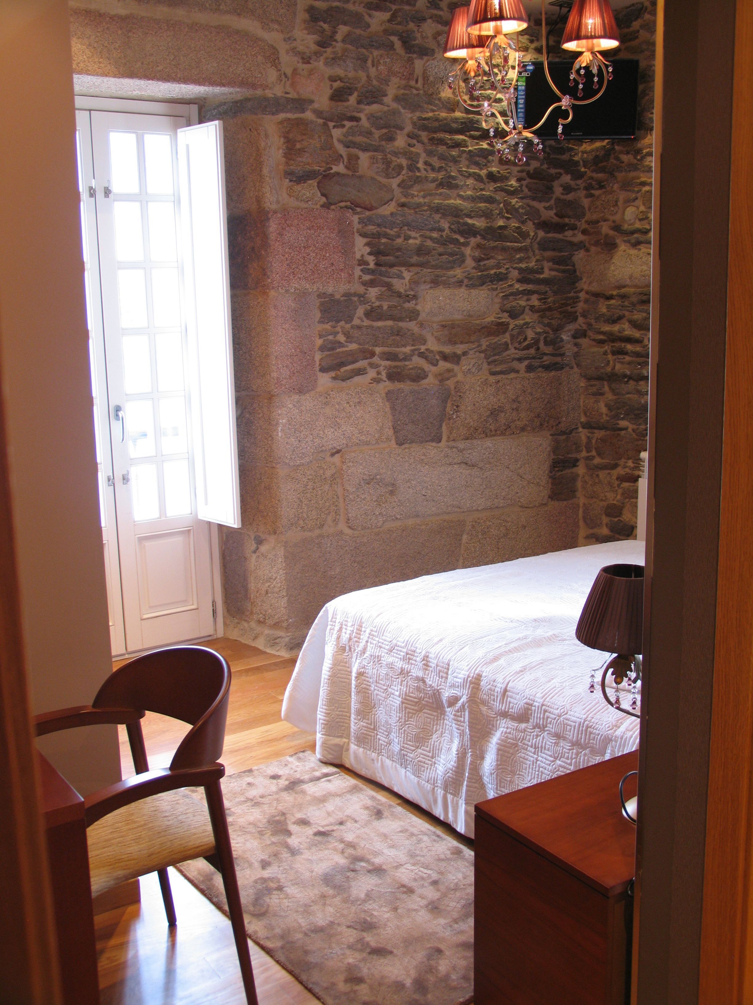 Un dormitorio con pared de piedra, una cama con cubrecama blanco, una silla de madera y un escritorio con lámpara.