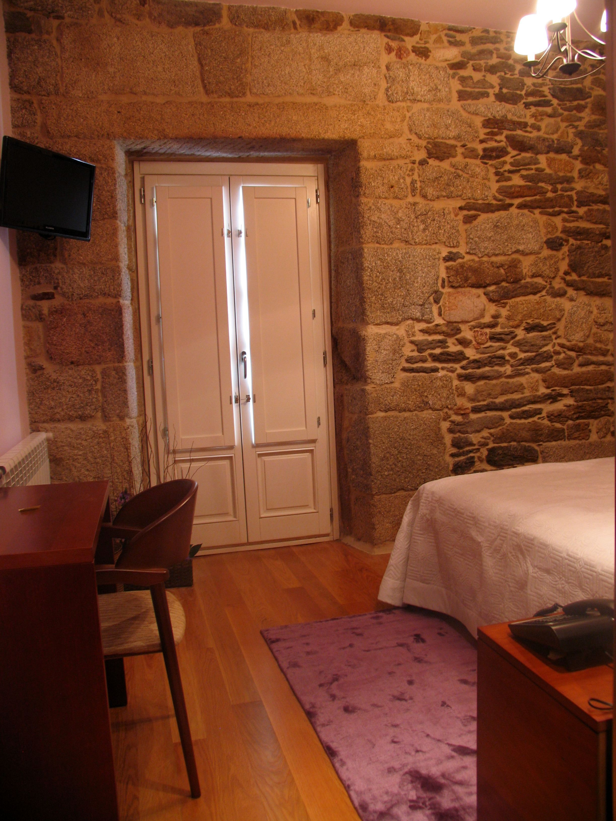 A room with a stone wall, a bed with a white bedspread, a desk with a chair, and a flat-screen TV mounted on the wall.