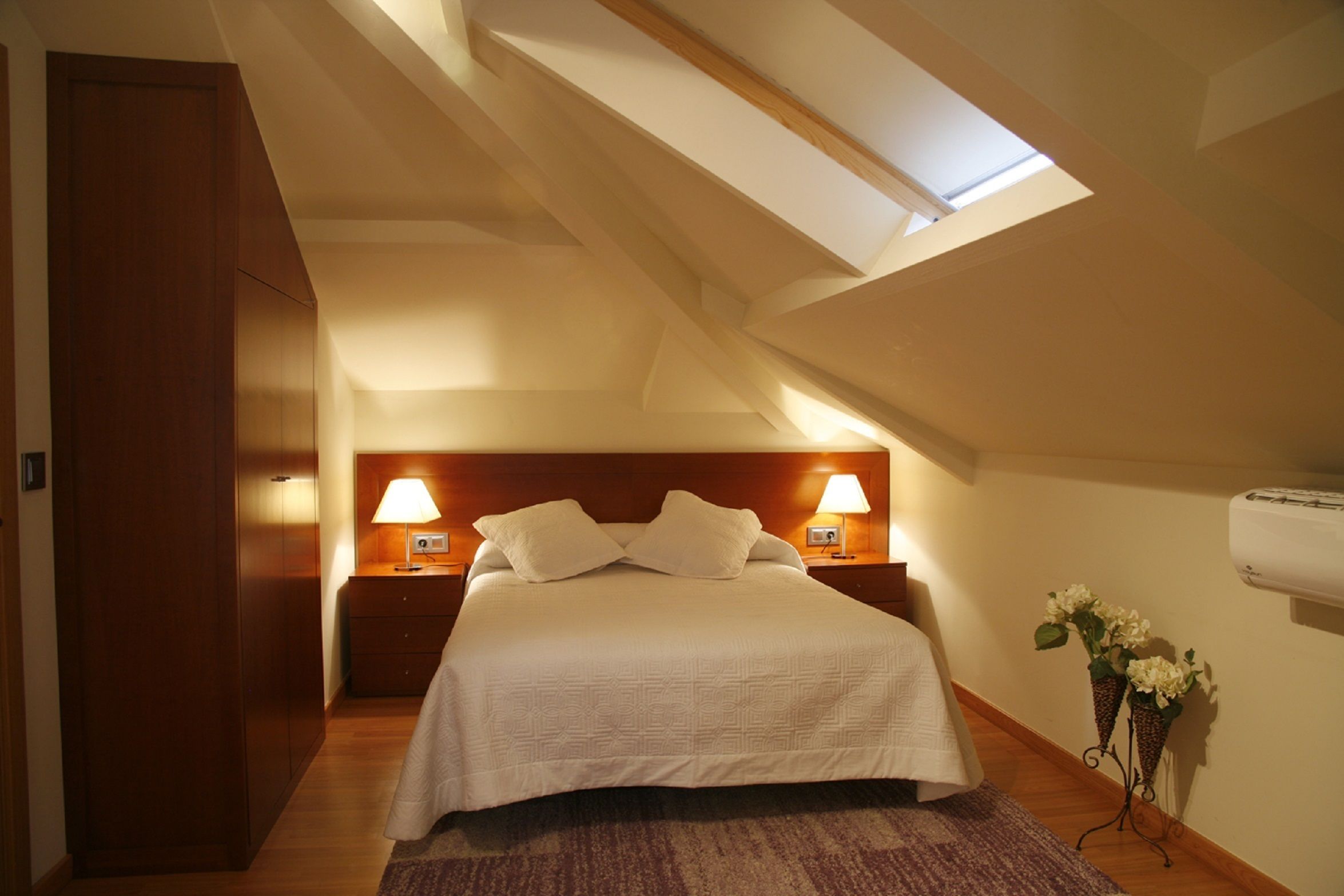 A bedroom with a sloped ceiling, a bed with white bedding, two bedside lamps, and a wall-mounted air conditioner.