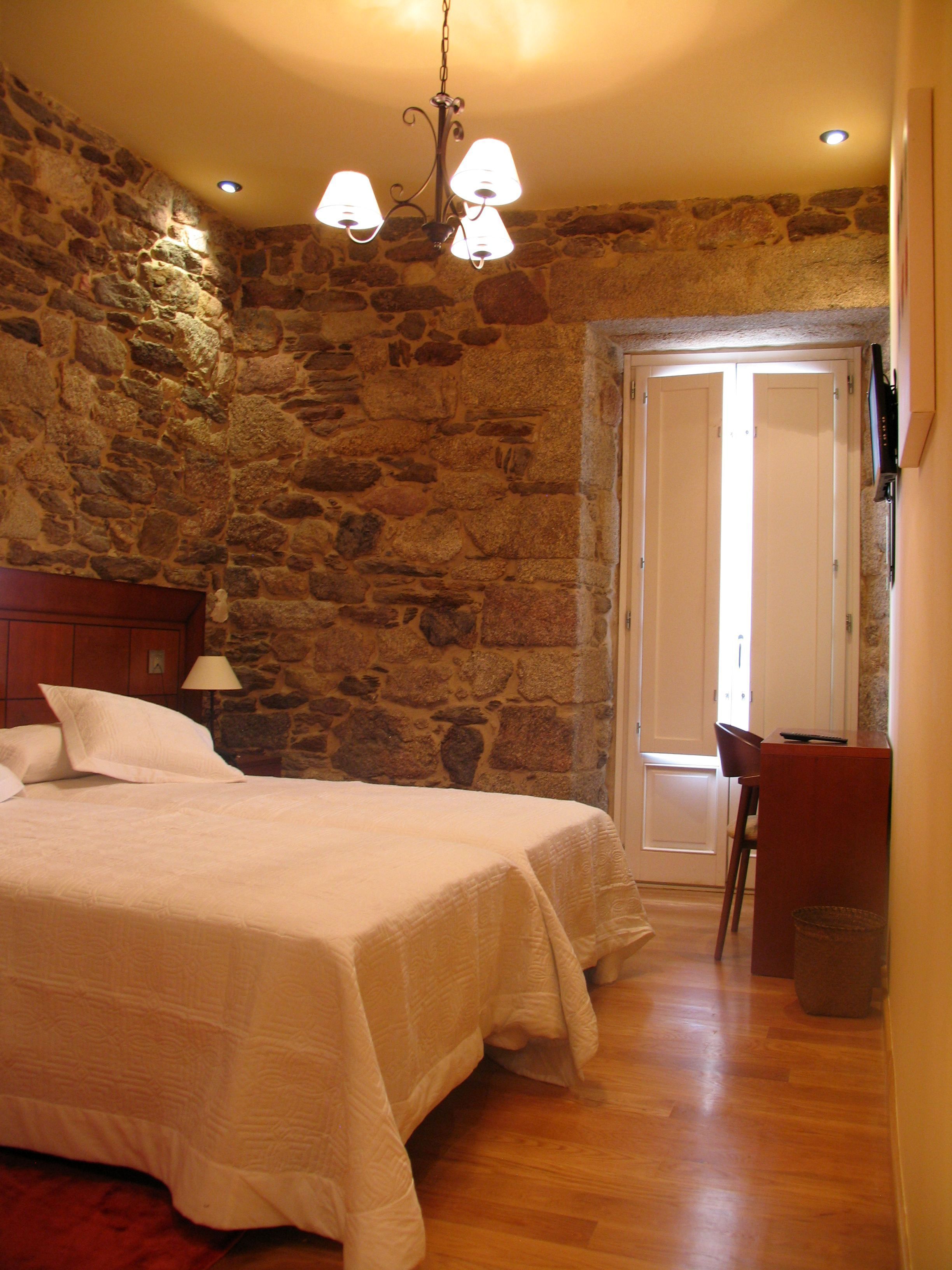 Un dormitorio con pared de piedra, cama con ropa de cama blanca, mesita de noche con lámpara y una silla de madera.