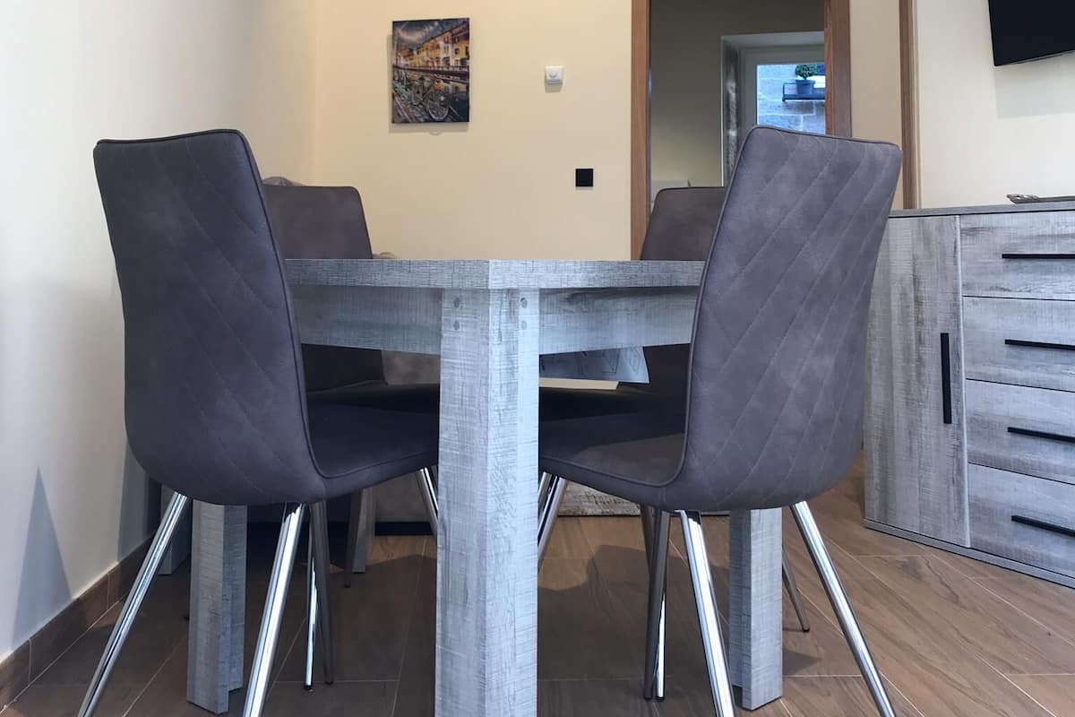 Uma sala de jantar com uma mesa de madeira e quatro cadeiras, um quadro na parede e uma televisão fixada na parede.