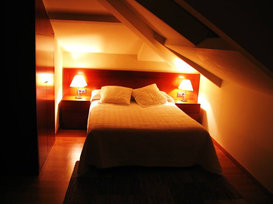 A dimly lit hotel room with a single bed, two bedside tables with lamps, and a sloped ceiling.