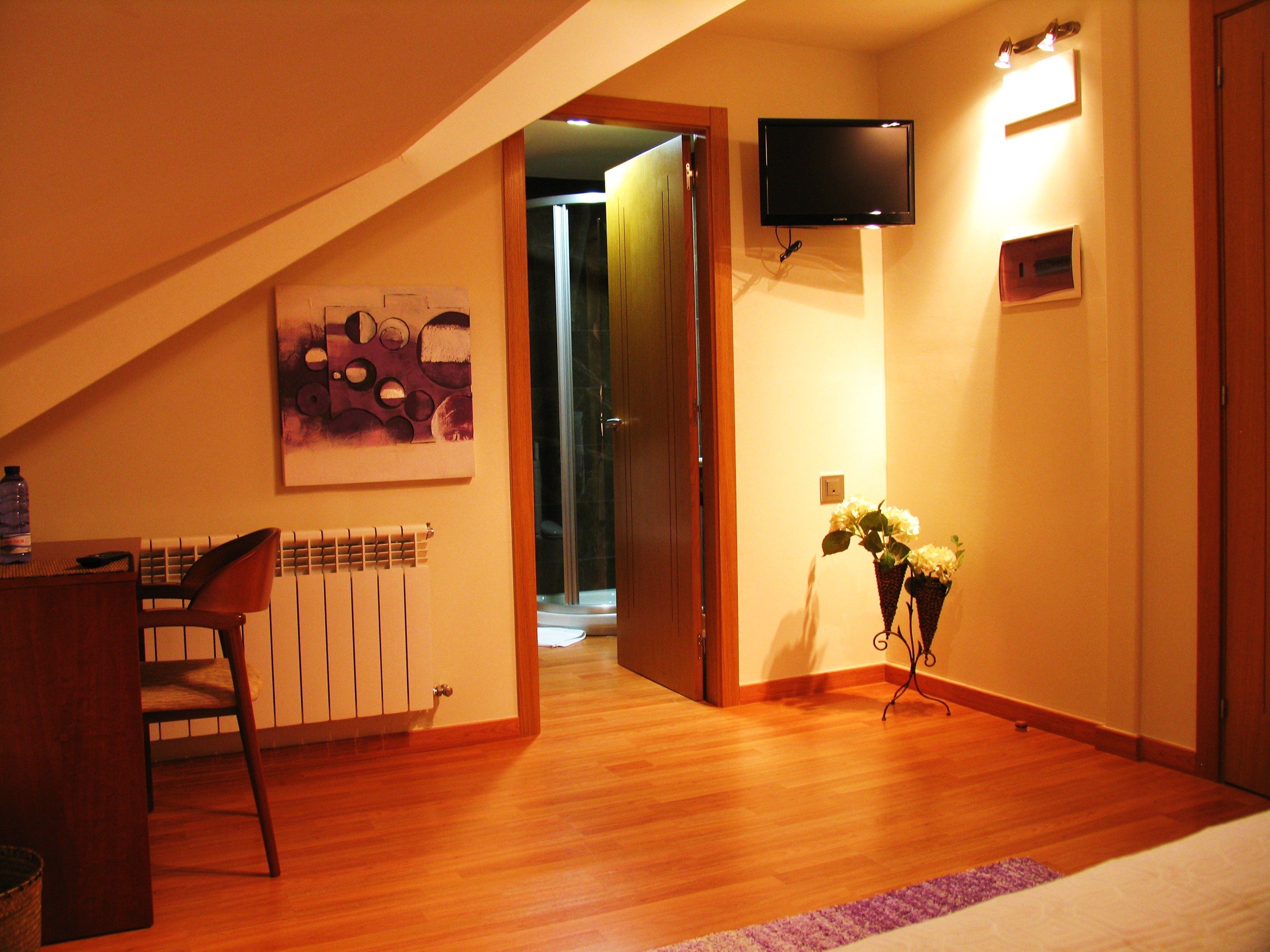 Een slaapkamer met een schuine kap, een bed, een bureau met stoel, een radiator, een aan de muur bevestigde televisie en een deur naar een andere kamer.