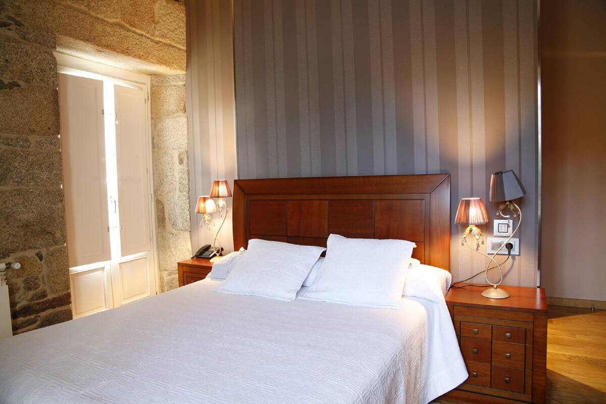 Cama bem arrumada com lençóis brancos num quarto com paredes de pedra e cabeceira de madeira. Há duas luminárias de cabeceira e uma mesa de cabeceira com abajur.