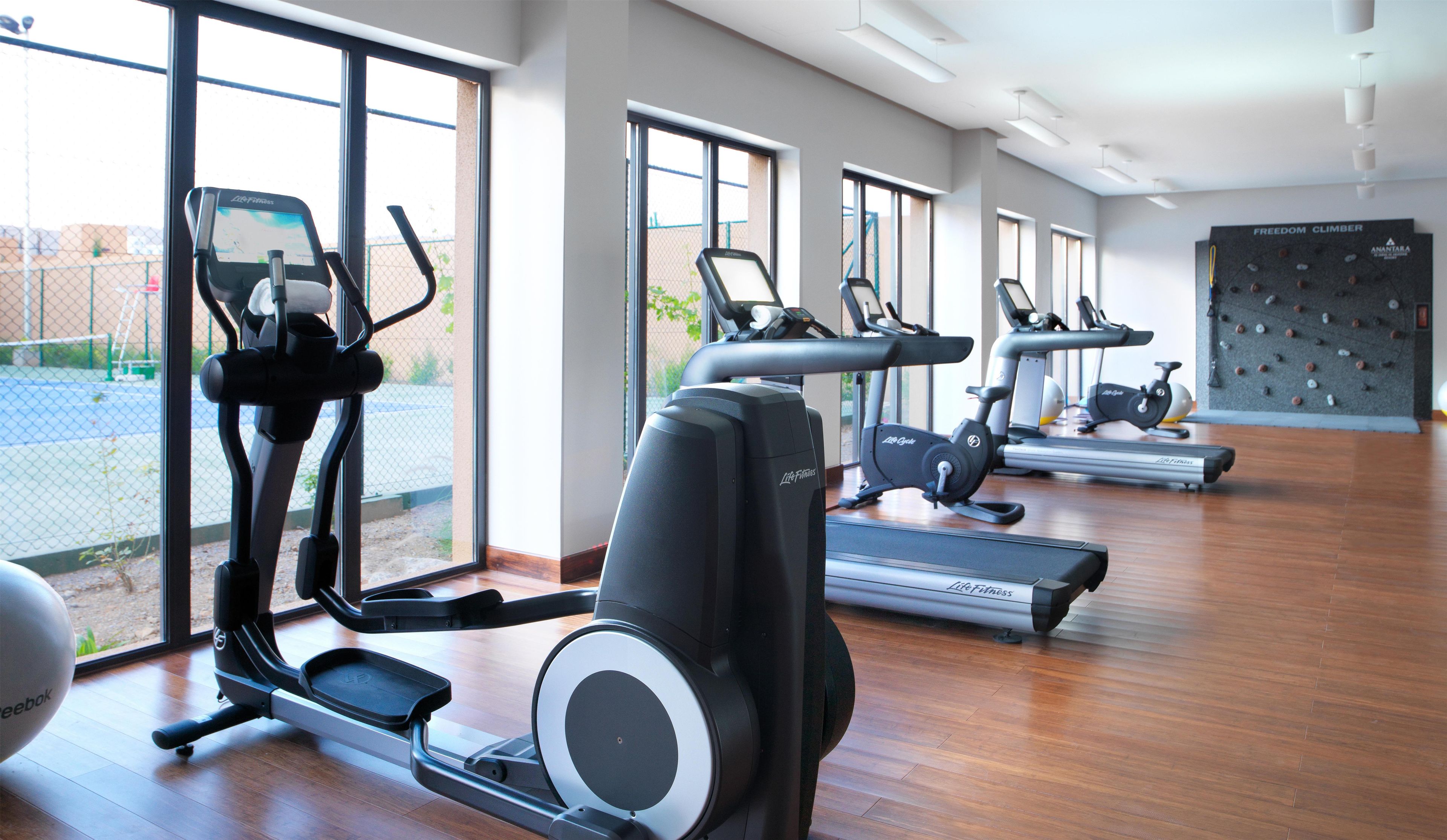 A gym with various cardio machines, large windows, and a wooden floor.