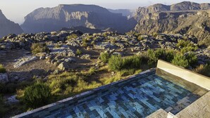 Una piscina con decorazioni in mosaico, circondata da terreno roccioso e vegetazione rada, con vista su un paesaggio montuoso.