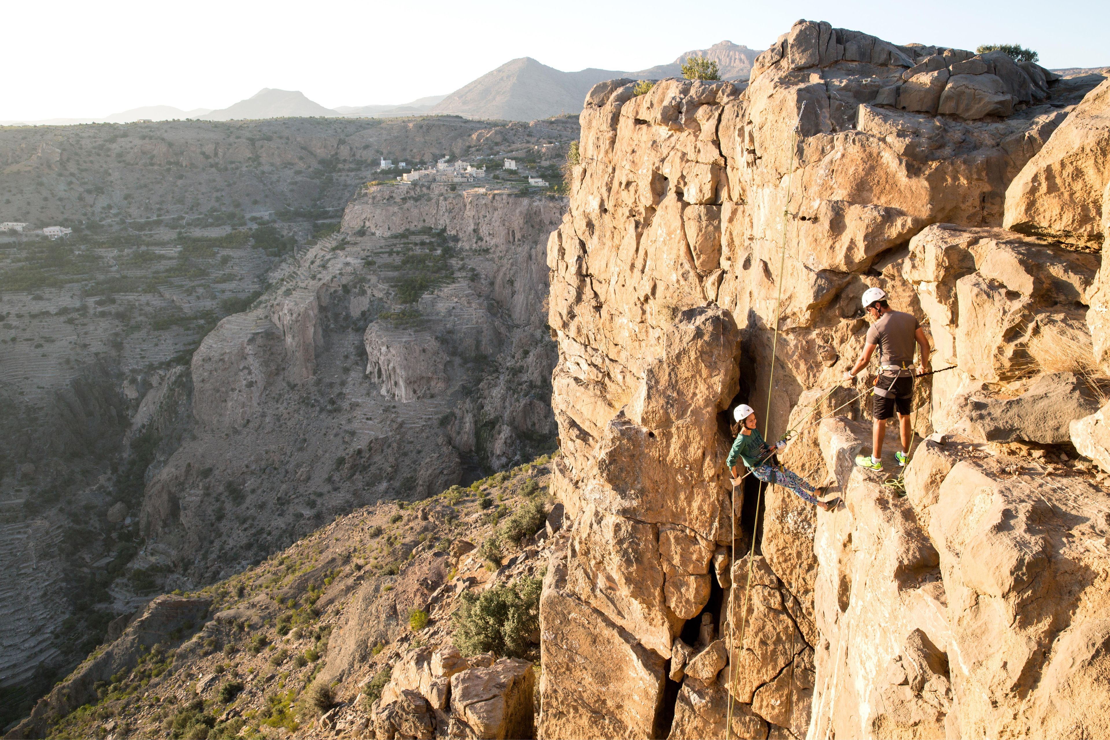 A rock climbing scene with a climber on a cliff, a second climber assisting, and a valley with buildings in the background.