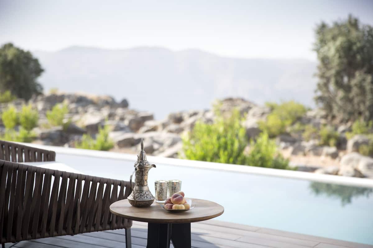 A poolside area with a wooden deck, a round table with a metal pitcher and glasses, and a plate of fruit.