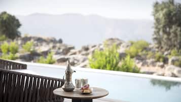 Un espace aménagé au bord de la piscine, avec une terrasse en bois, une table ronde, une carafe et des verres en métal, ainsi qu’une assiette de fruits.