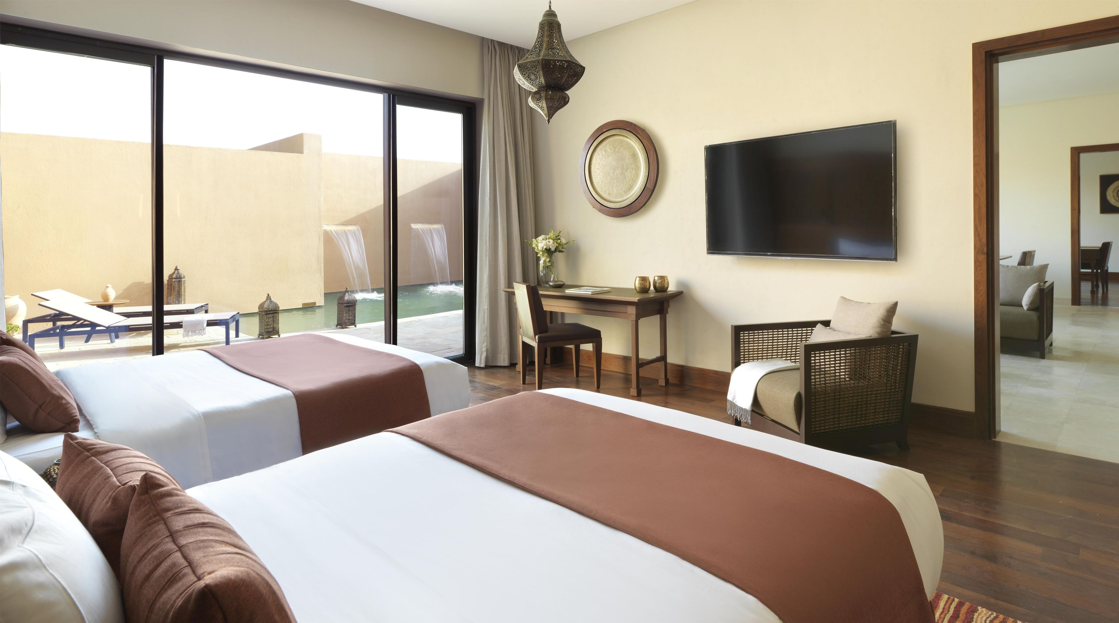 A hotel room with two beds, a desk, a chair, a TV, and a view of a pool.