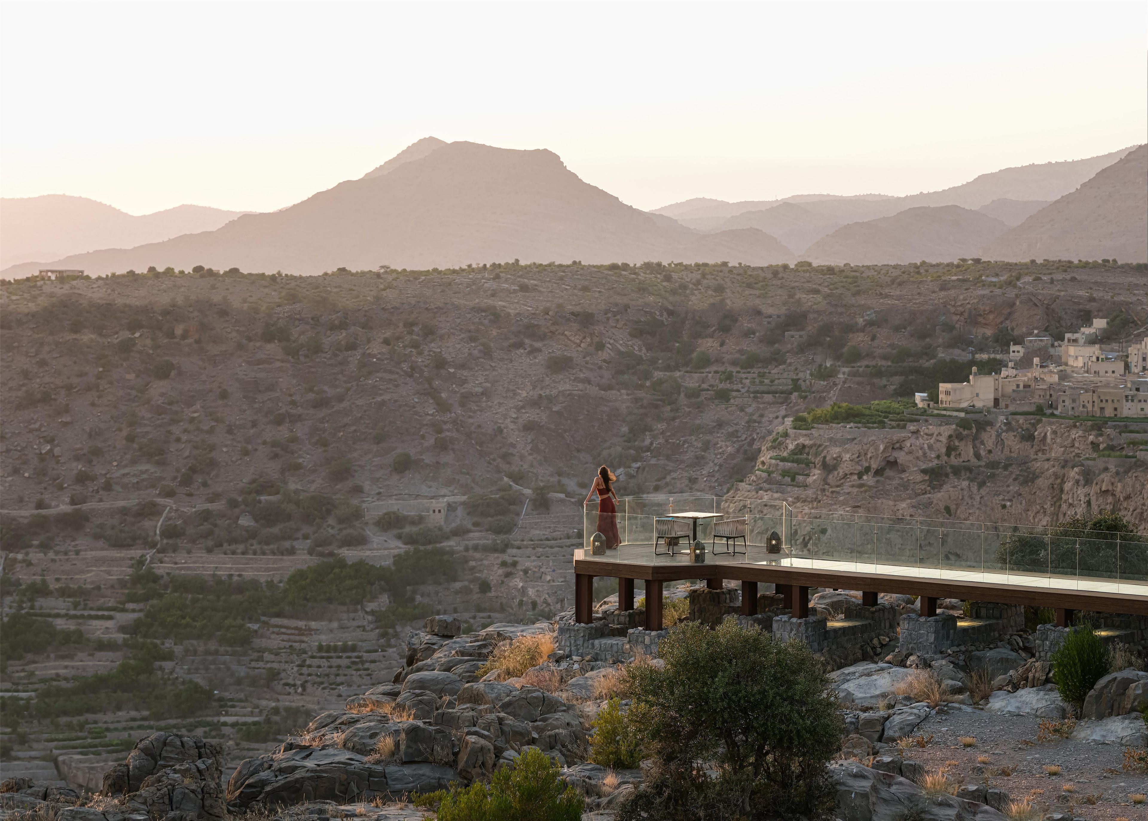 A glass-roofed terrace with a table and chairs, overlooking a mountainous landscape with terraced fields and a village.